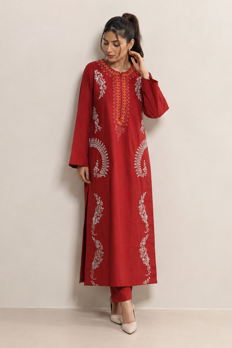A woman modeling a red long kurta with white and orange embroidery, paired with matching red pants and white heels, standing against a plain beige wall.