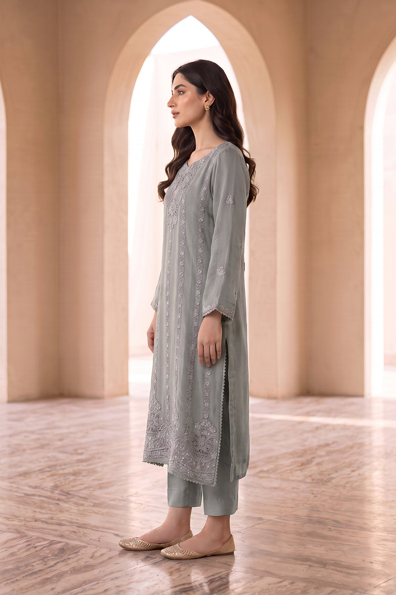 A woman stands in profile wearing a light sage green embroidered kurta with matching pants, set against a warm beige architectural background with arches.