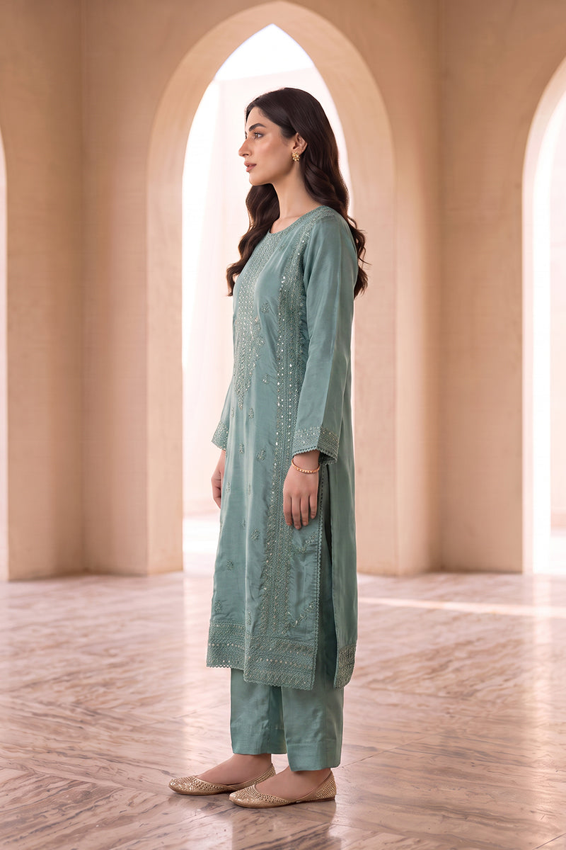 A woman stands in profile wearing a sage green embroidered kurta and matching pants set, posed in a warm, sunlit architectural hallway with arched doorways.