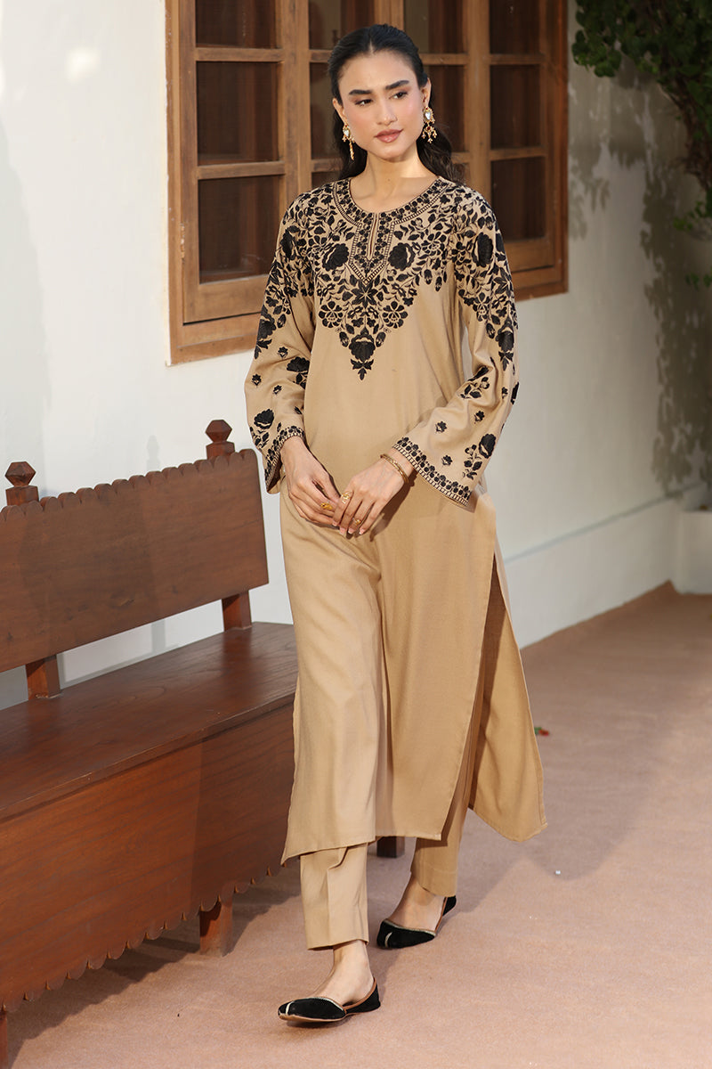 A woman models a beige and black embroidered traditional outfit with matching trousers, standing in front of a white wall with wooden window frames and a wooden bench.
