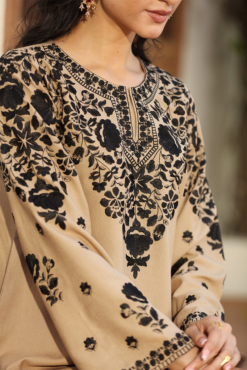 A close-up of a woman wearing a beige long-sleeve tunic or kurta with prominent black floral embroidery and geometric border detailing around the neckline and sleeves.