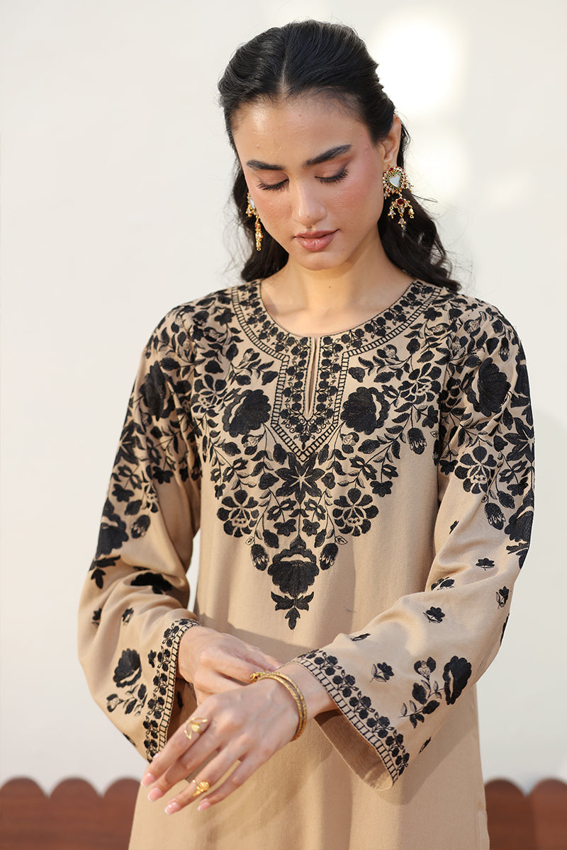 A woman wearing a beige long-sleeve tunic with black floral embroidery and geometric patterns, styled with gold jewelry against a light neutral background.