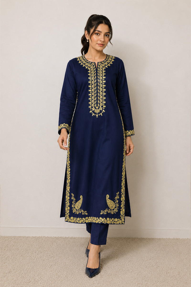 A woman modeling a navy blue traditional kurta set with ornate gold embroidery around the neckline, cuffs, hem, and peacock motifs near the lower front, paired with matching navy pants and dark pointed shoes against a plain light wall.