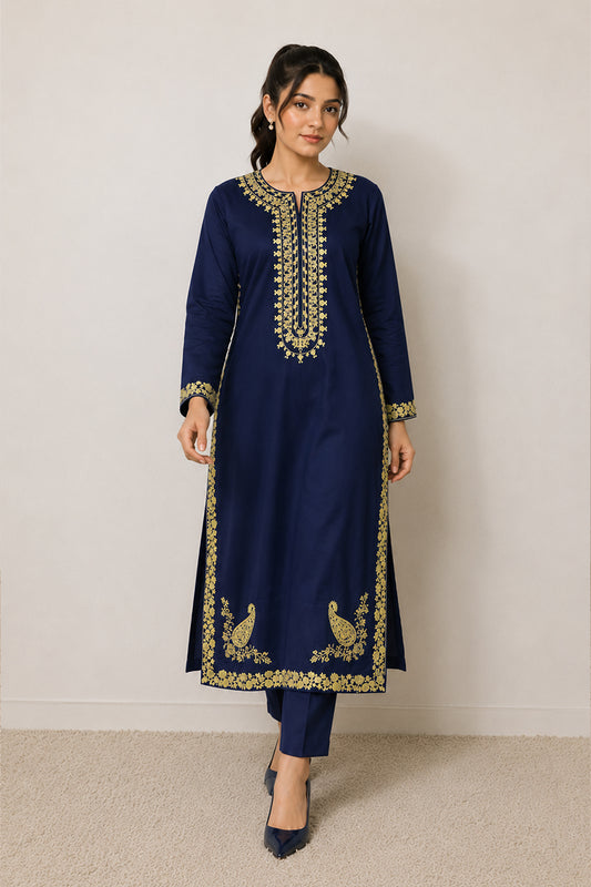 A woman modeling a navy blue traditional kurta set with ornate gold embroidery around the neckline, cuffs, hem, and peacock motifs near the lower front, paired with matching navy pants and dark pointed shoes against a plain light wall.