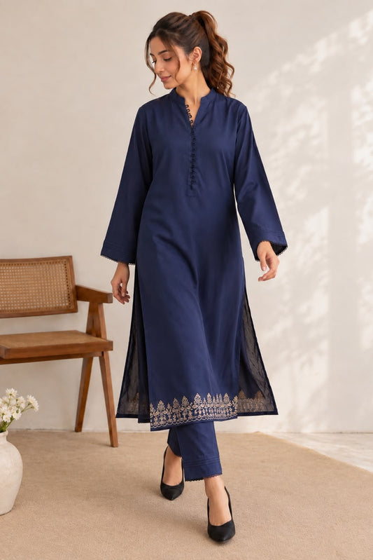 A woman modeling a navy blue kurta set with matching trousers in a bright, modern room. The kurta has a mandarin collar, buttoned placket, and a decorative embroidered border near the hem.