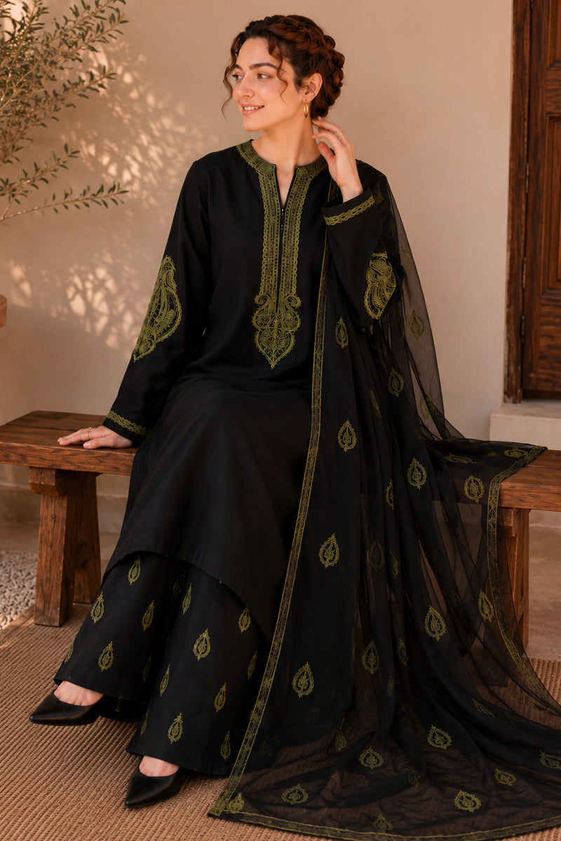 A woman models a black three-piece traditional outfit with olive green embroidery, consisting of a long kurta, matching trousers, and a sheer dupatta, seated on a rustic wooden bench.