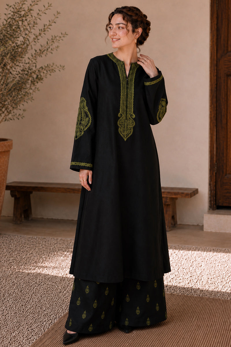A woman modeling a black long kurta with green embroidered details on the neckline, sleeves, and hem, paired with matching black wide-leg pants with green motifs, set in a rustic indoor environment.