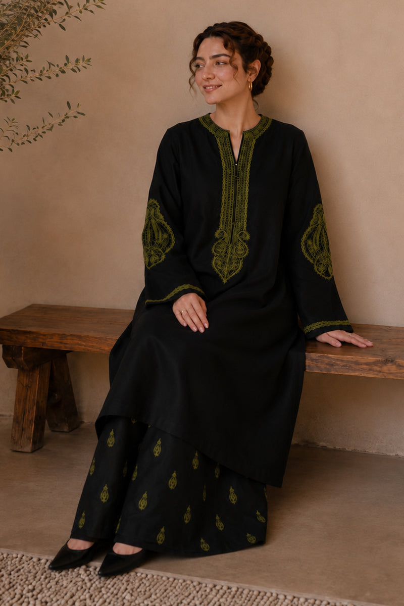 A woman wearing a black traditional kurta set with olive green embroidery sits on a rustic wooden bench against a beige stucco wall.