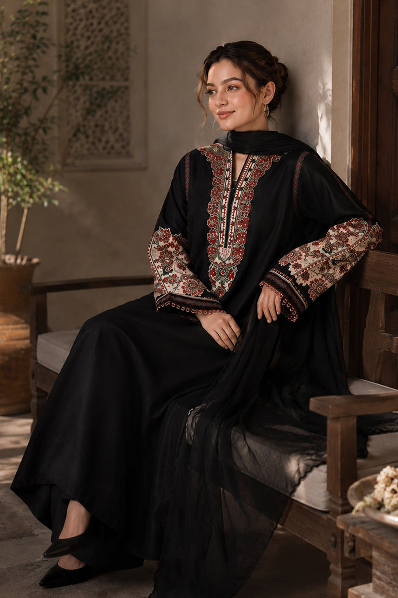 A woman seated on a wooden bench wearing a black traditional outfit with ornate multicolor embroidered detailing on the neckline and sleeves, in a softly lit indoor setting.