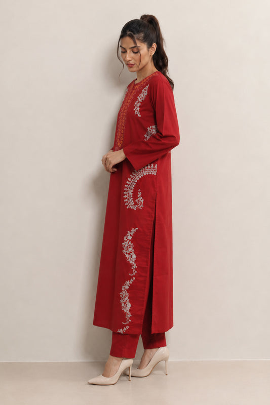 A woman modeling a long red kurta with white embroidered patterns and matching red trousers, standing against a plain light beige wall.