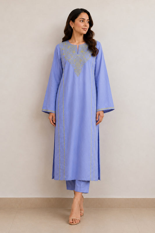 A woman modeling a light blue straight kurta with gold embroidery on the chest and along the front, paired with matching blue pants, set against a plain beige wall.