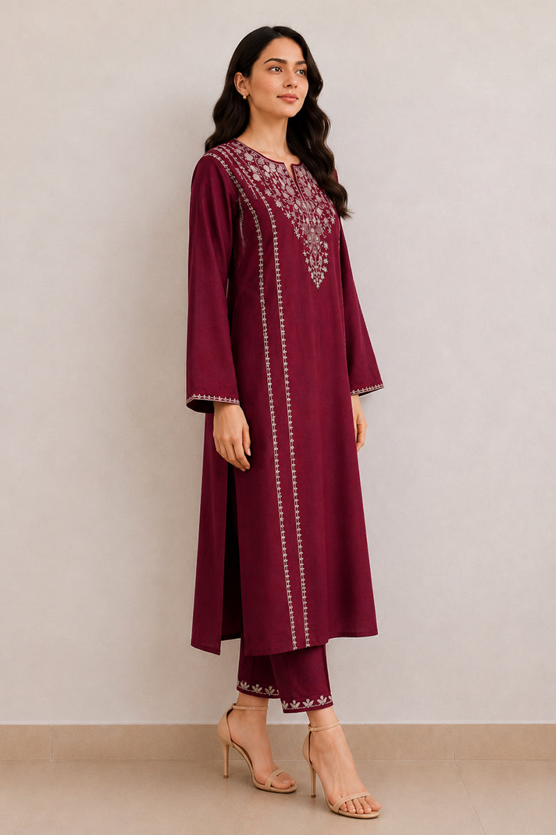 A woman modeling a deep maroon three-piece kurta set with silver mirror work embroidery on the neckline and vertical borders, paired with matching pants and nude high-heeled sandals, set against a plain beige studio background.