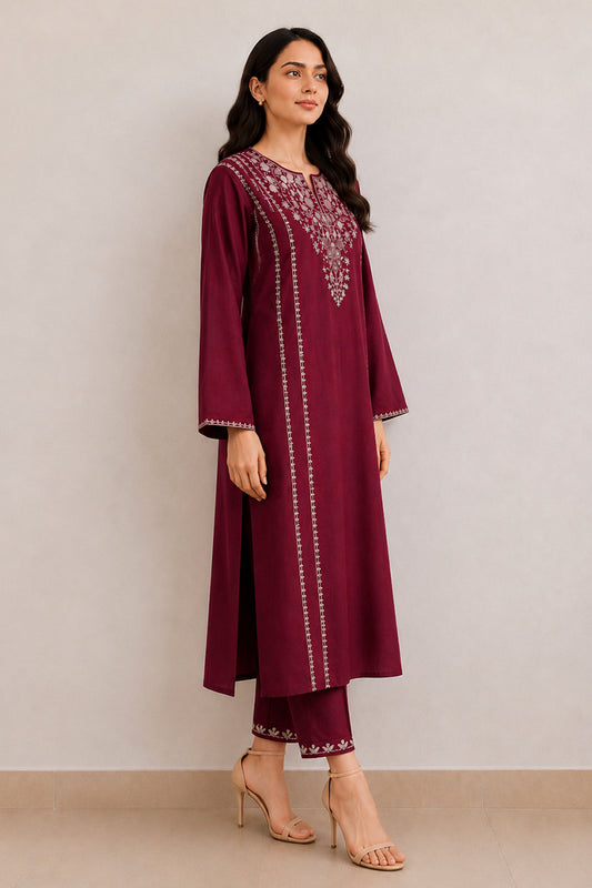 A woman modeling a deep maroon three-piece kurta set with silver mirror work embroidery on the neckline and vertical borders, paired with matching pants and nude high-heeled sandals, set against a plain beige studio background.