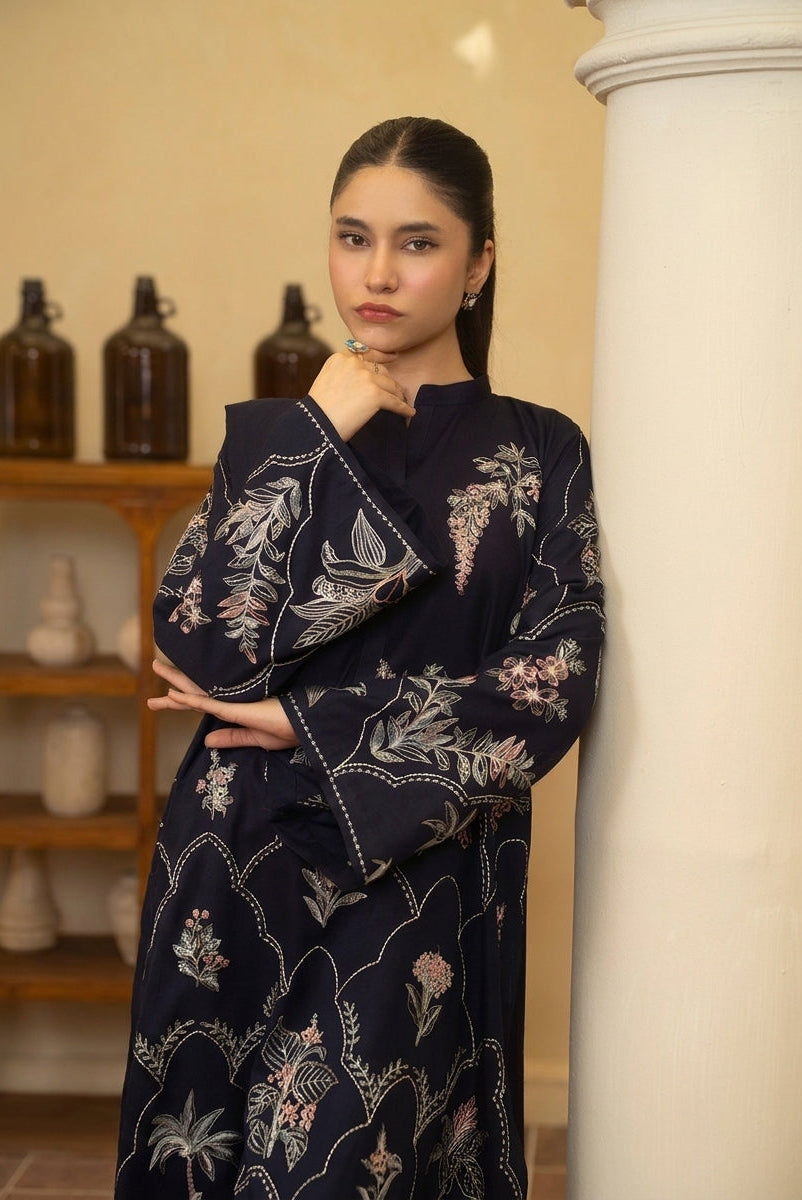 A woman wearing a dark navy blue long-sleeved dress with white and pink floral embroidery, posing indoors next to a white column.