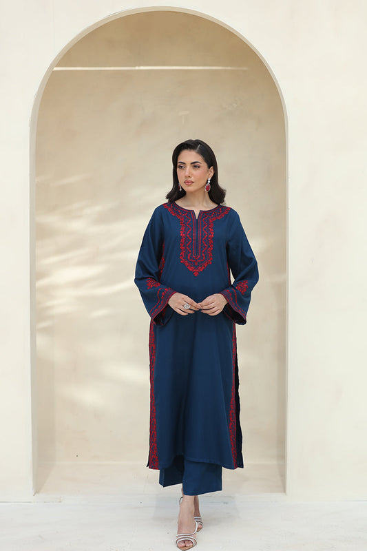 A woman modeling a navy blue kurta with red embroidered detailing on the neckline and sleeve cuffs, paired with matching navy pants, standing in front of a beige arched wall.
