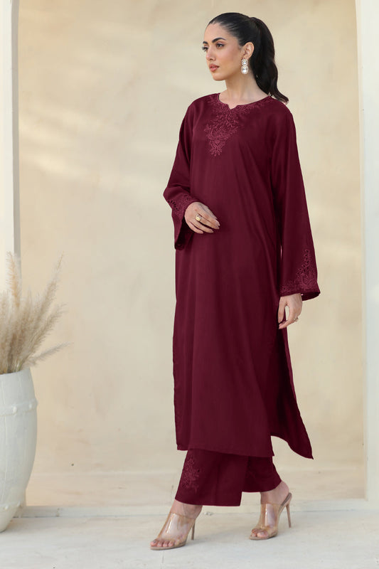 A woman models a three-piece maroon kurta set with matching pants and clear high-heeled sandals, standing against a beige wall with a decorative vase on the left.