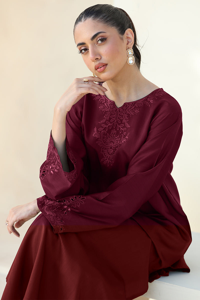 A woman wearing a long-sleeved maroon dress with floral embroidery on the chest and cuffs, posed against a neutral beige background.