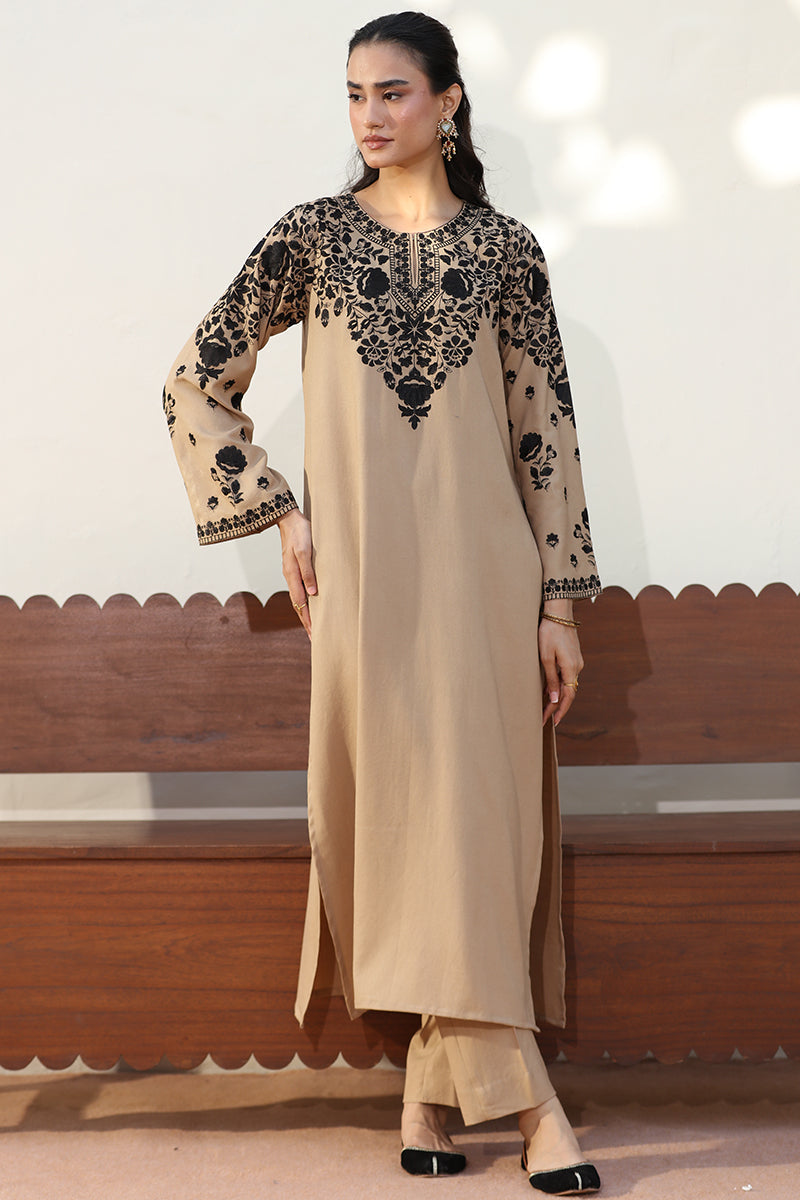 A woman models a beige long kurta with black floral embroidery on the yoke and sleeves, paired with matching beige trousers, standing in front of a wooden bench.