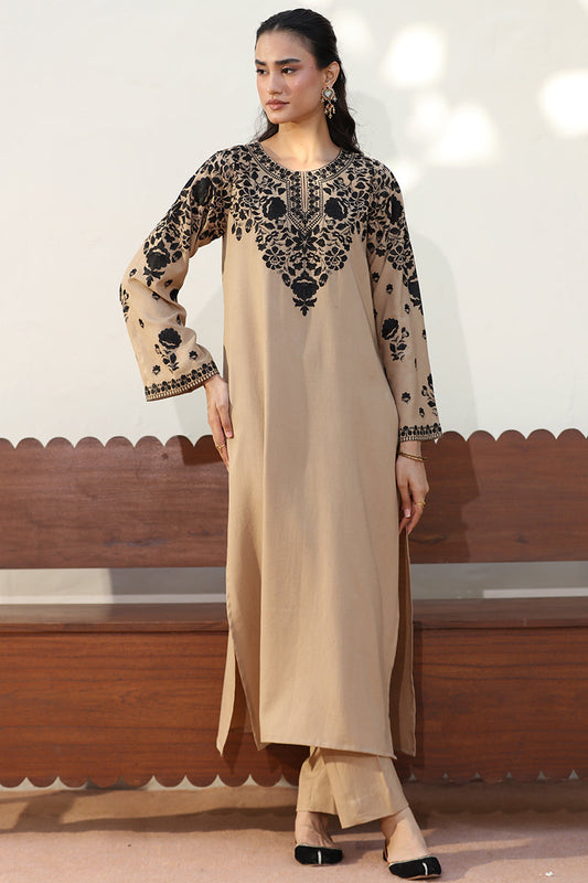 A woman models a beige long kurta with black floral embroidery on the yoke and sleeves, paired with matching beige trousers, standing in front of a wooden bench.