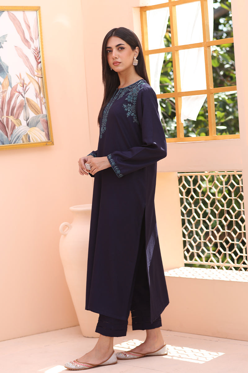A woman models a dark navy blue long kurta with teal embroidery on the neckline and cuffs, paired with matching dark cropped pants, in a warm peach-colored room with a window and decorative wall art.