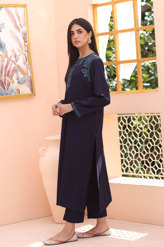 A woman models a dark navy blue long kurta with teal embroidery on the neckline and cuffs, paired with matching dark cropped pants, in a warm peach-colored room with a window and decorative wall art.