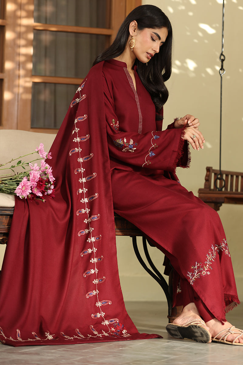 A woman seated on a wooden bench wearing a deep maroon traditional outfit with embroidered details and a matching draped scarf. The setting appears to be an indoor porch or balcony with warm neutral walls and window light.