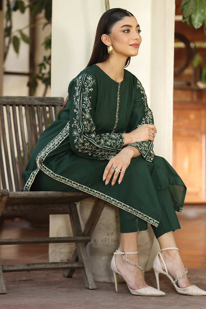 Woman in a green embroidered outfit sitting on a wooden bench.