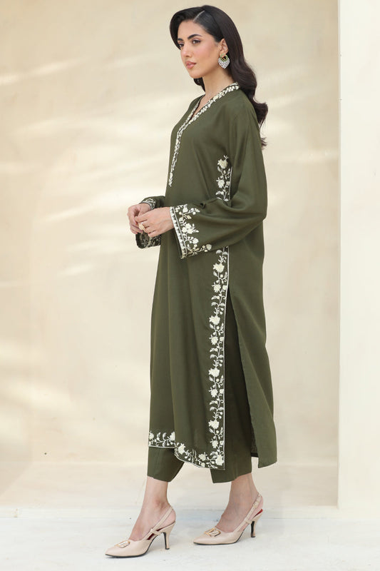 A woman models an olive green long kurta with white floral embroidery on the neckline, cuffs, and side panels, paired with matching olive green trousers and beige high-heeled mule sandals, set against a warm beige studio backdrop.
