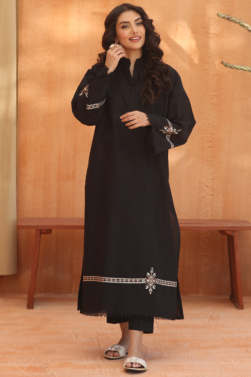 A woman modeling a long black tunic with silver and white embroidered details on the sleeves and hem, standing in front of a warm brown textured wall.