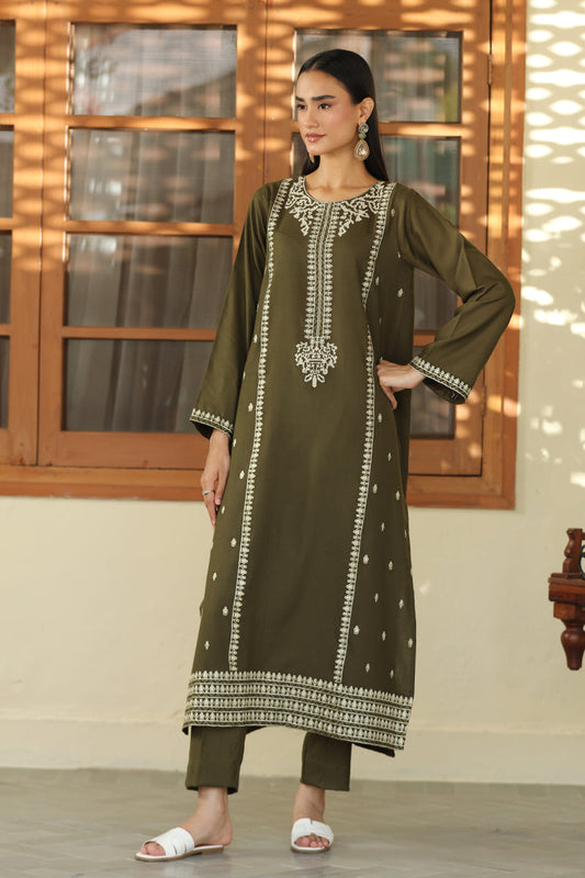 A woman models an olive green long kurta with intricate silver mirror work embroidery on the neckline, front panel, cuffs, and hem, paired with matching olive green cropped pants and white slide sandals, set against a warm-toned wooden window background.