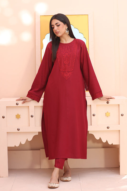 A woman models a long-sleeved maroon red kurta with matching trousers, featuring embroidered detailing on the chest, posed in front of a light pink cabinet.