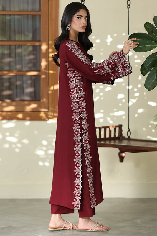 A woman modeling a long maroon kurta with intricate white floral embroidery on the front and sleeves, paired with matching maroon pants, in a styled indoor setting with a hanging swing and green leaves.
