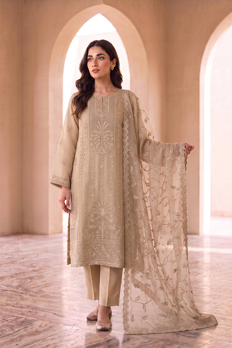 A woman models a traditional beige three-piece salwar kameez suit with intricate embroidery, featuring a long kurta, matching trousers, and a sheer embroidered dupatta, set against an elegant arched corridor.
