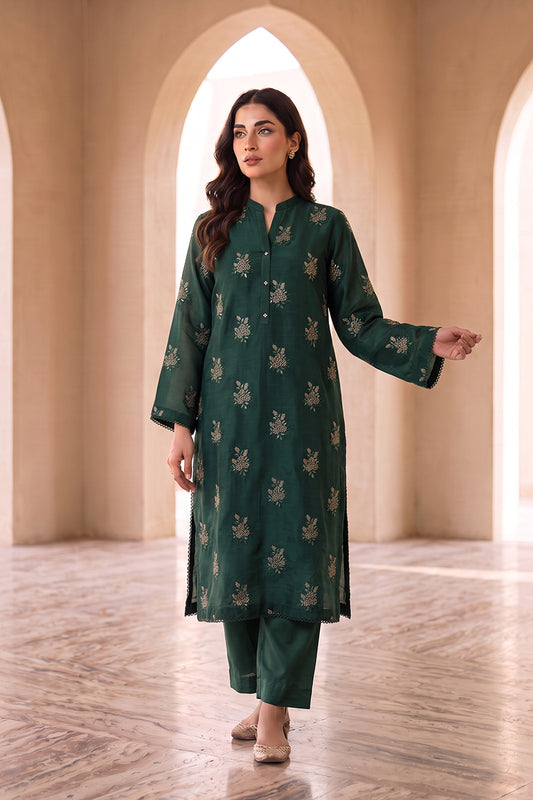 A woman modeling a dark green three-piece kurta set with beige floral embroidery, standing in a grand hall with beige arches and marble floors.