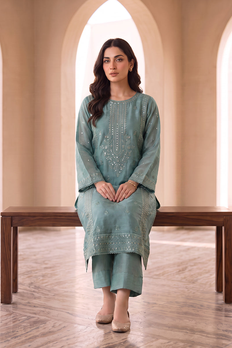 A woman models a sage green three-piece traditional outfit with embroidered details, consisting of a long kurta, pants, and a dupatta, seated on a wooden bench in an arched room.