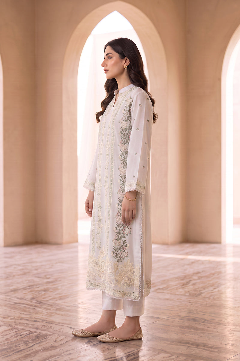 A woman stands in profile wearing a long white embroidered kurta with matching white pants and beige flat shoes, posed in a warm architectural setting with arched windows and marble flooring.
