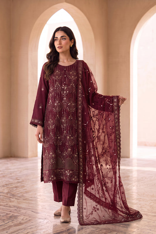 A woman models a three-piece traditional outfit in deep maroon, featuring a long kurta with intricate gold embroidery, matching trousers, and a sheer embroidered dupatta, set against a background of beige arched columns.