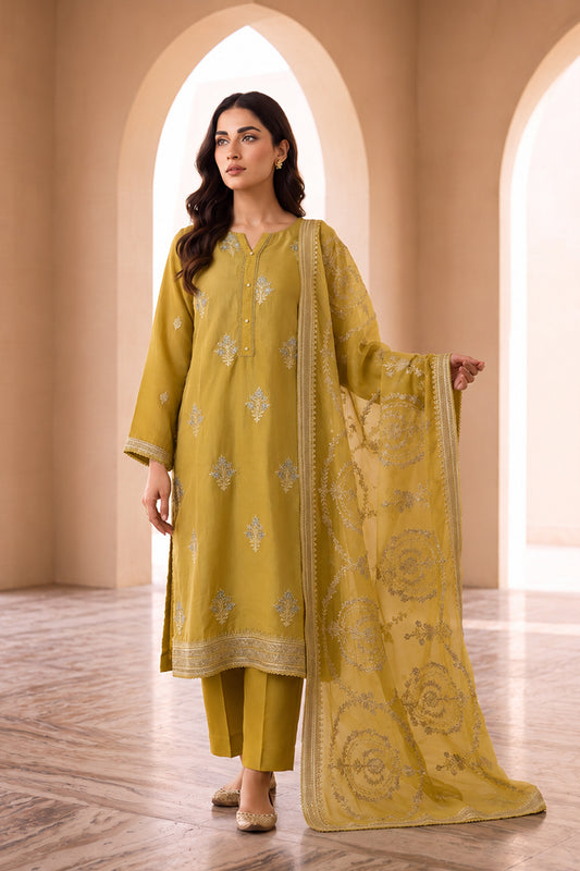 A woman models a three-piece mustard yellow ethnic suit set featuring a kurta with silver embroidery, matching trousers, and a sheer dupatta with floral prints, set against a warm beige arched background.