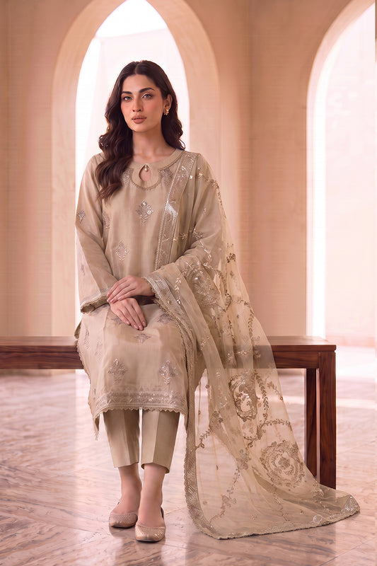 A woman models a traditional beige three-piece salwar kameez suit with matching dupatta in a warm, arched interior setting. The set features a beige cotton kameez kurta with silver embroidery, matching beige trousers, and a sheer beige dupatta with gold floral embroidery.