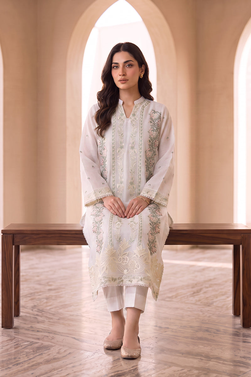 A woman models a traditional white embroidered kurta set with matching white trousers, standing in front of a wooden bench inside an arched room with beige walls.
