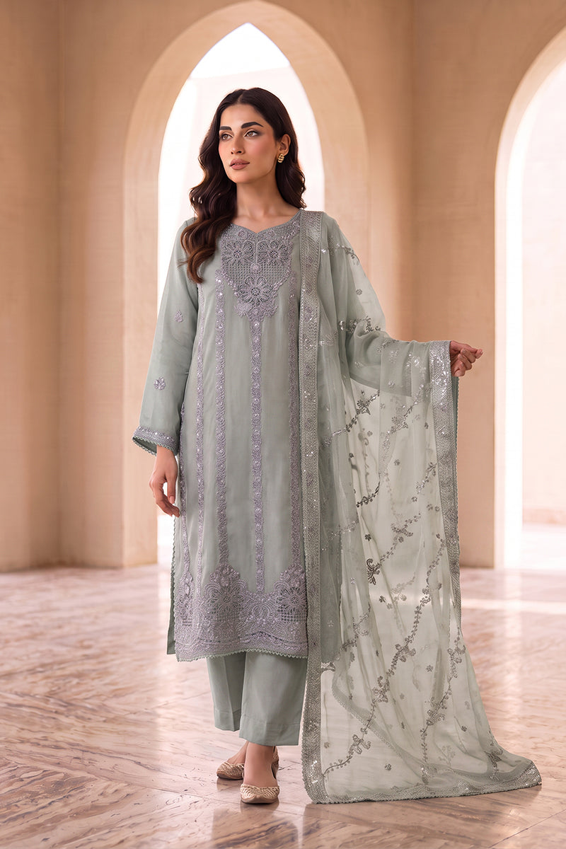 A woman models a light sage green traditional Indian outfit with matching pants and a sheer embroidered dupatta, set against a warm beige architectural background with arches.