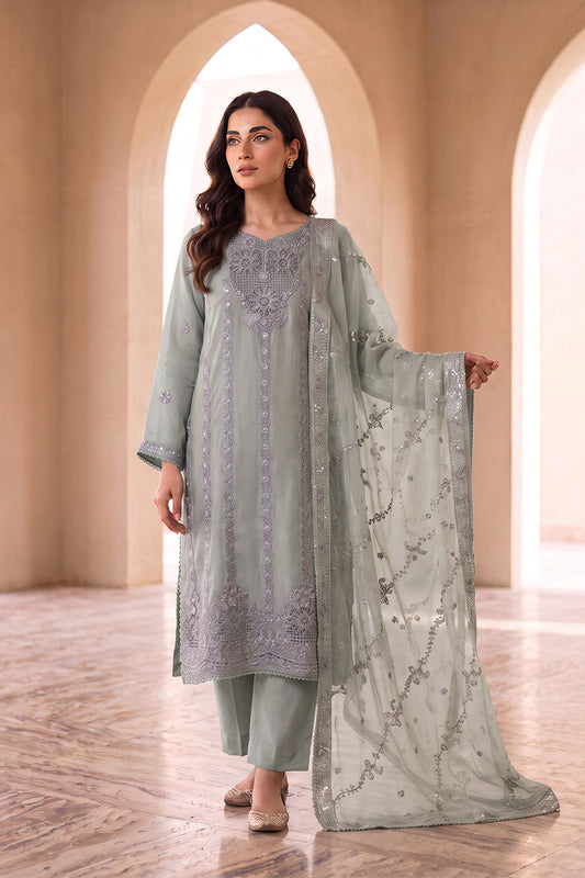 A woman models a light sage green traditional Indian outfit with matching pants and a sheer embroidered dupatta, set against a warm beige architectural background with arches.