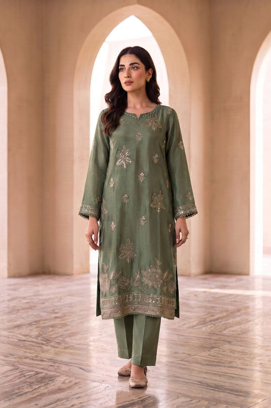 A woman modeling a sage green three-piece salwar kameez suit with beige floral embroidery, standing in a warm, arched corridor with stone walls and a marble floor.