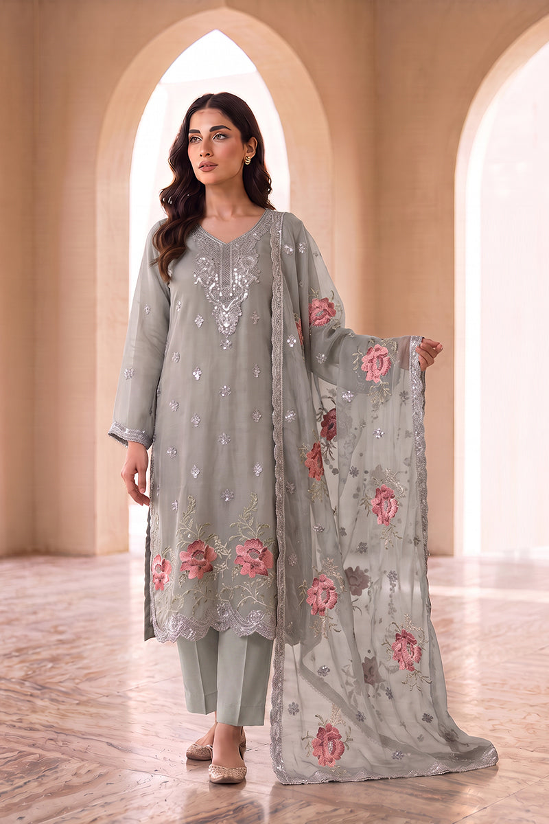 A woman models a traditional three-piece salwar kameez set in a warm, arched corridor. The set features a light gray-green kurta with silver mirror work embroidery, matching trousers, and a sheer dupatta with pink floral embroidery.