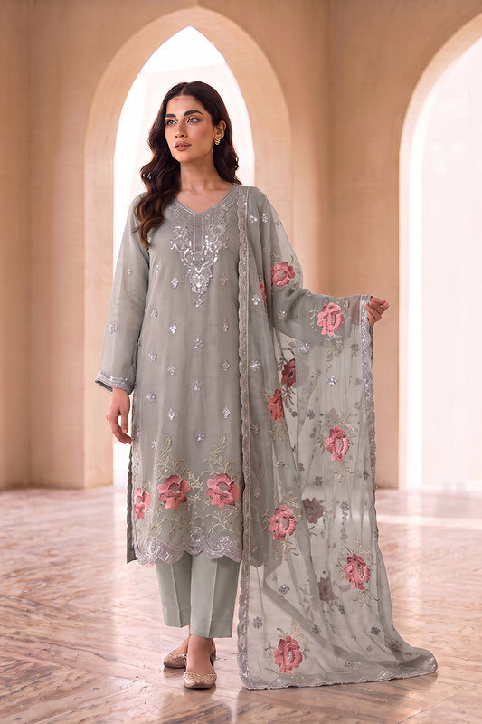 A woman models a traditional three-piece salwar kameez set in a warm, arched corridor. The set features a light gray-green kurta with silver mirror work embroidery, matching trousers, and a sheer dupatta with pink floral embroidery.