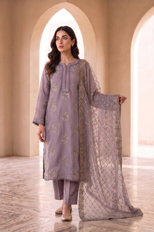 A woman models a three-piece traditional outfit in a warm beige arched corridor. The outfit is a muted lavender-gray color with gold floral embroidery on the kurta and matching trousers, and includes a sheer embroidered dupatta.