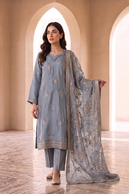A woman models a traditional three-piece salwar kameez suit in a warm beige arched corridor. The suit is light blue-gray with intricate gold and silver floral embroidery, paired with matching trousers and a sheer dupatta.