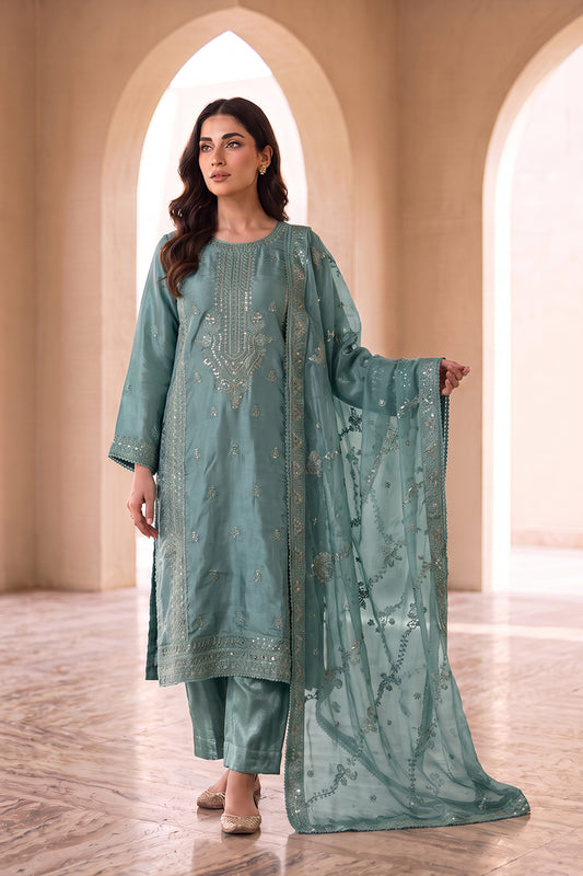 A woman models a three-piece traditional outfit in a dusty sage green color with silver embroidery, consisting of a long kurta, matching trousers, and a sheer dupatta, set against a background of beige arched columns.