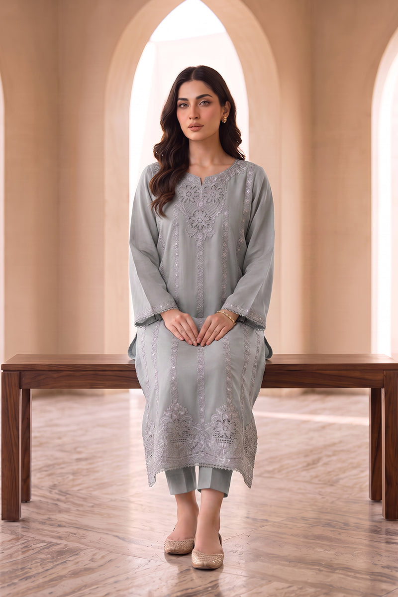 A woman models a light gray three-piece salwar kameez set with intricate silver embroidery, standing in front of a wooden bench in a warm, arched interior.